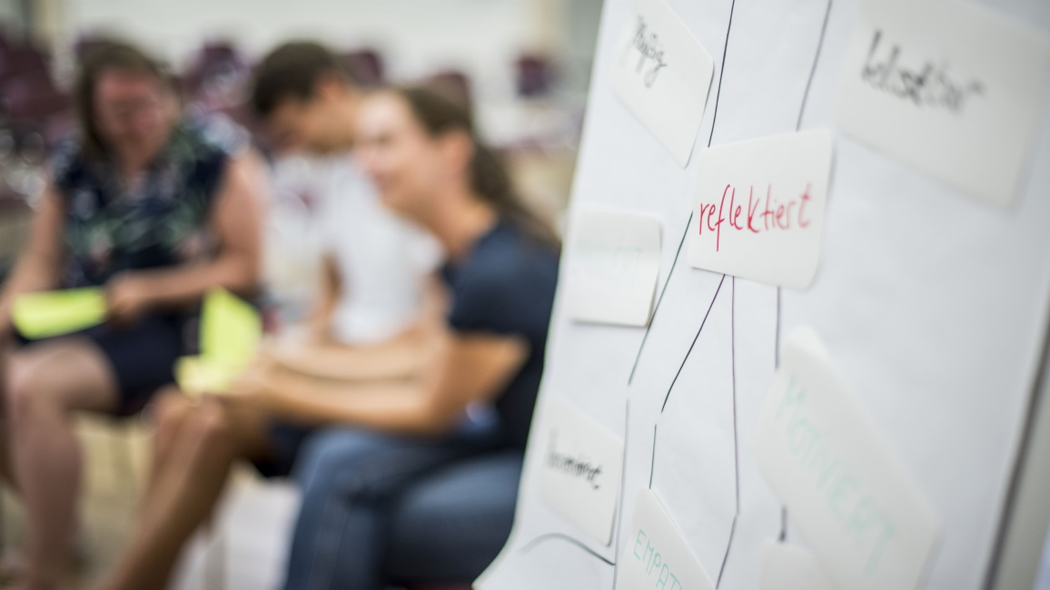 Mehrere Personen sitzen in einem Raum zusammen. Im Vordergrund ist ein Flipchart zu sehen.