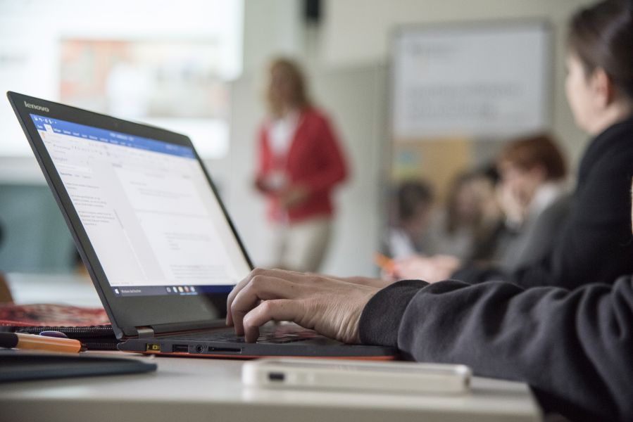 Eine Person sitzt an einem Tisch und tippt auf der Tastatur eines Laptops. Im Hintergrund steht eine Person.