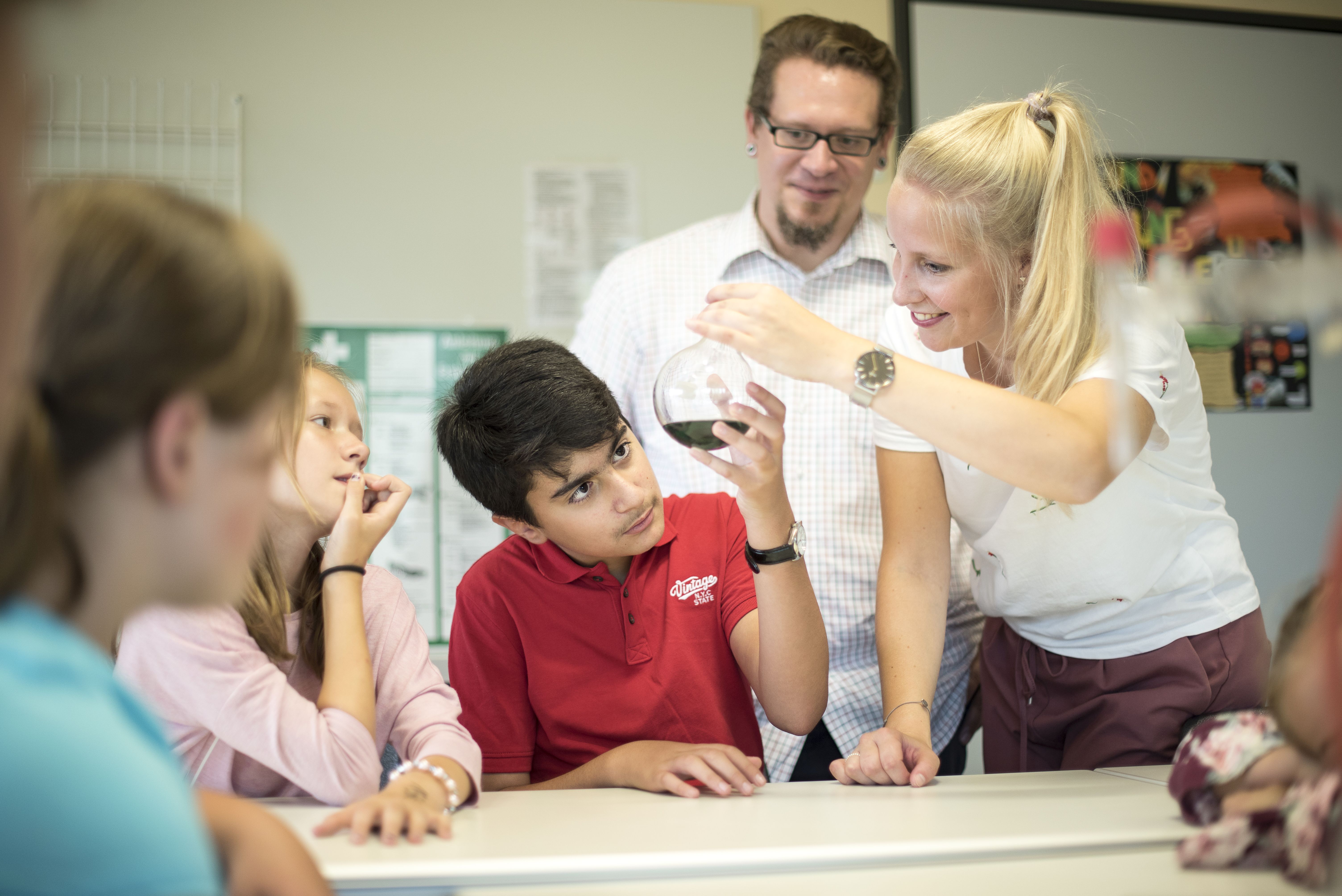 Lernsituation mit Lehrkräften und Schülerinnen und Schülern. Frau zeigt einem Jungen ein Laborglas.