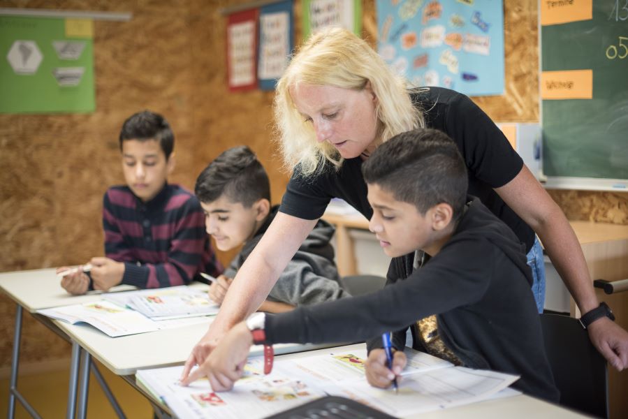 Drei Schüler sitzen im Klassenraum. Eine Lehrerin beugt sich zu einem Schüler und zeigt in ein Schulbuch