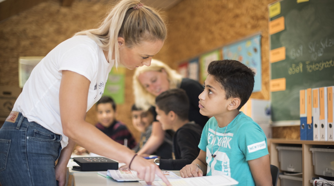 Eine Lehrerin zeigt in ein Buch und erklärt einem Schüler etwas. Er blickt zu ihr hinauf. Im Hintergrund sind weitere Schüler und eine Lehrerin zu sehen. 