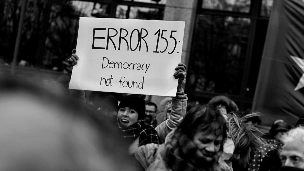 Schwarzweißfoto einer Demonstration: Eine junge Frau hält ein Plakat mit der Aufschrift „ERROR 155: Democracy not found” hoch. 