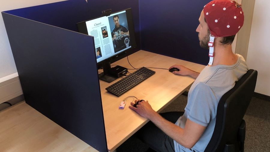Ein Mann sitzt vor einem Computerbildschirm und einer Tastatur in einer Kabine und hält in der rechten Hand eine Maus. An den Fingern der linken Hand befinden sich Sensoren, zudem trägt er eine Haube mit Sensoren auf dem Kopf. 