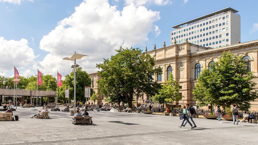 Auf einem großen Platz vor einem historischen Gebäude sitzen junge Menschen auf Bänkenund laufen 