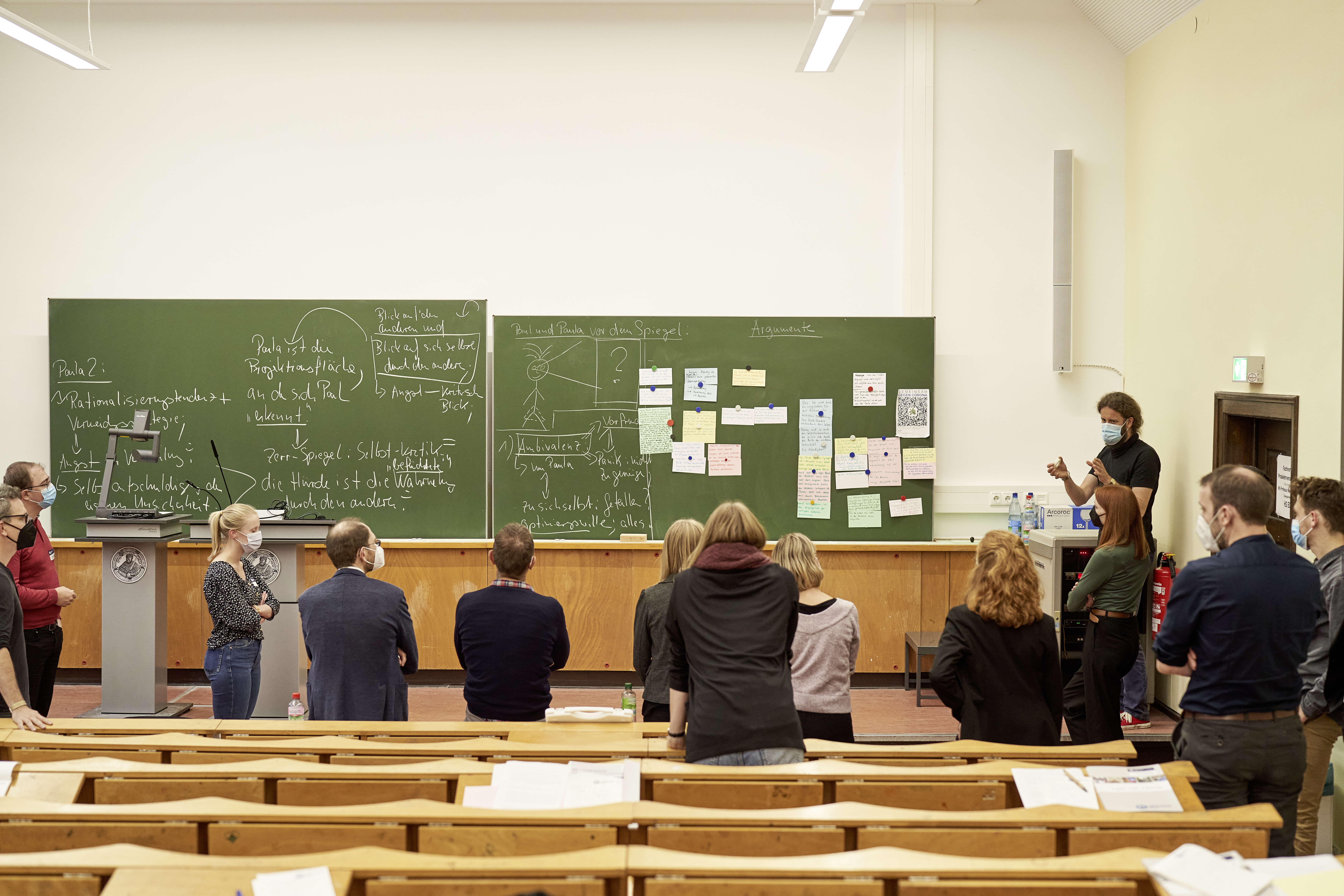 In einem Hörsaal stehen Leute vorne vor zwei großen Tafeln.