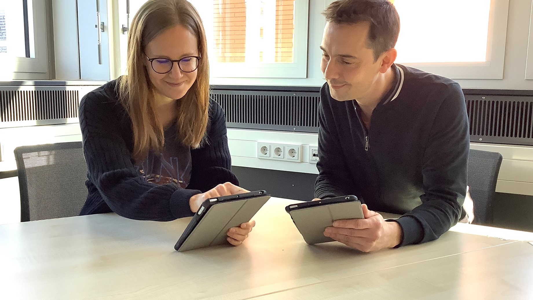 Eine junge Frau und ein junger Mann sitzen an einem Tisch, jeder hält ein Tablet in der Hand.