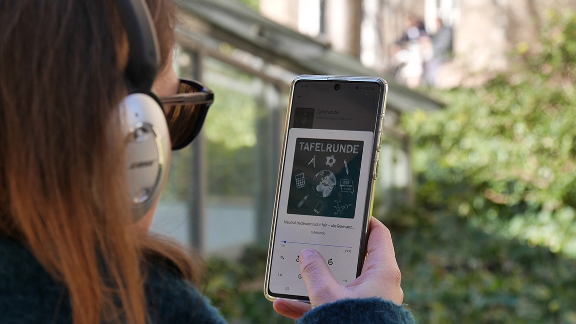 Eine junge Frau mit Sonnenbrille und Kopfhörer ist von hinten zu sehen. Sie hält in der rechten Hand ein Smartphone, dessen Screen das Logo des Podcasts „Tafelrunde“ der Heidelberg School of Education zeigt.