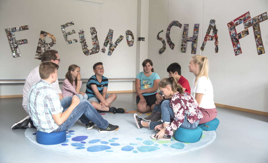 Ein Lehrer sitzt mit Schülern und Schülerinnen in einem Kreis. An der Wand steht groß das Wort Freundschaft.