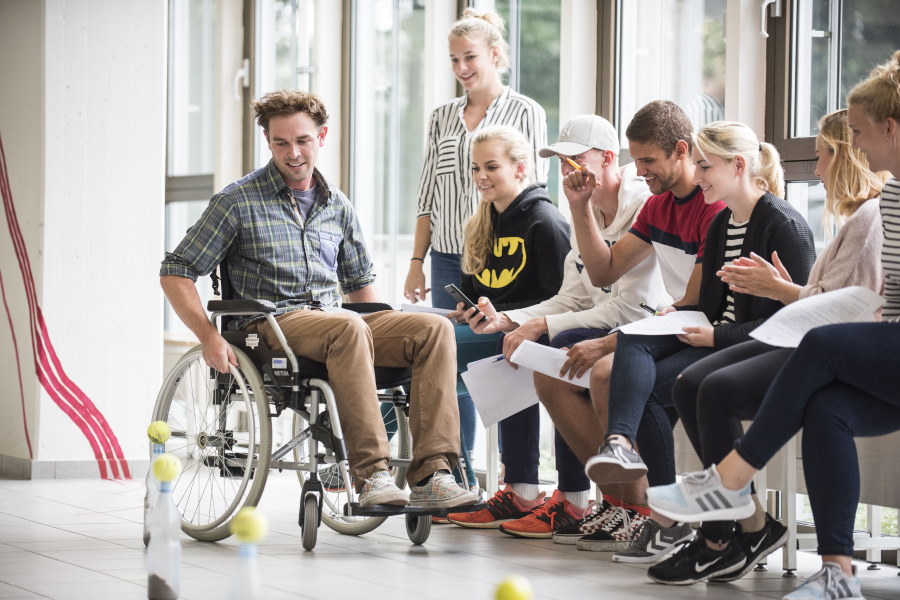 Ein junger Mann im Rollstuhl spielt ein Ballspiel, Zuschauer feuern ihn an