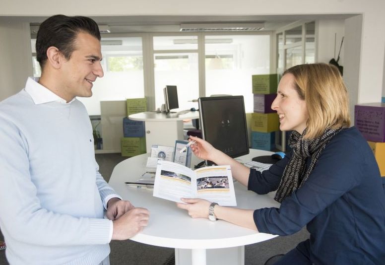 Ein Mann und eine Frau im Gespräch an einem Stehtisch, die Frau hält eine geöffnete Broschüre in der Hand.