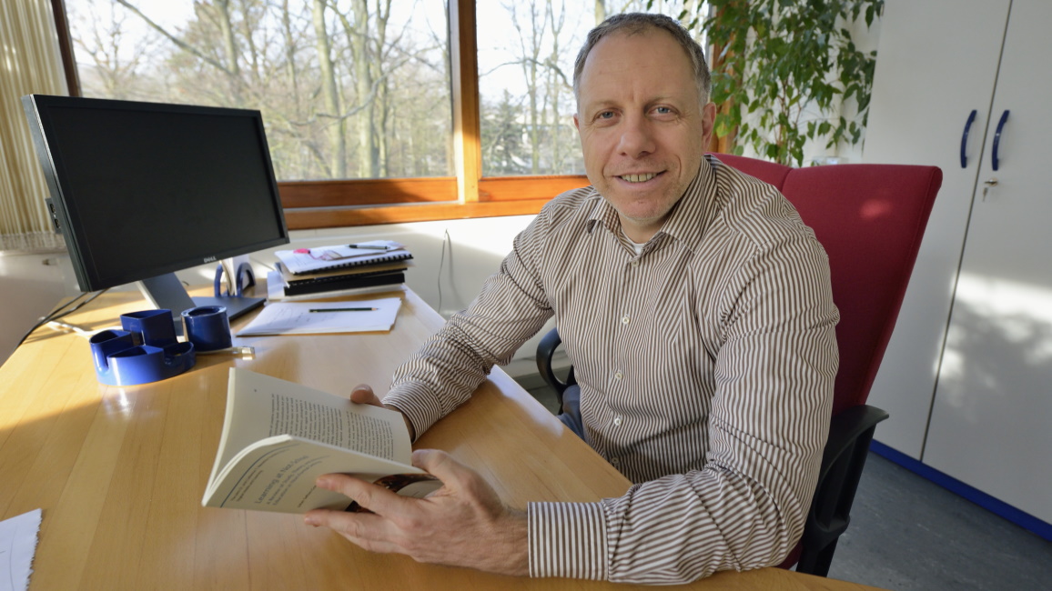 Professor Ludwig Stecher sitzt an einem Schreibtisch und hält ein Buch in der Hand.
