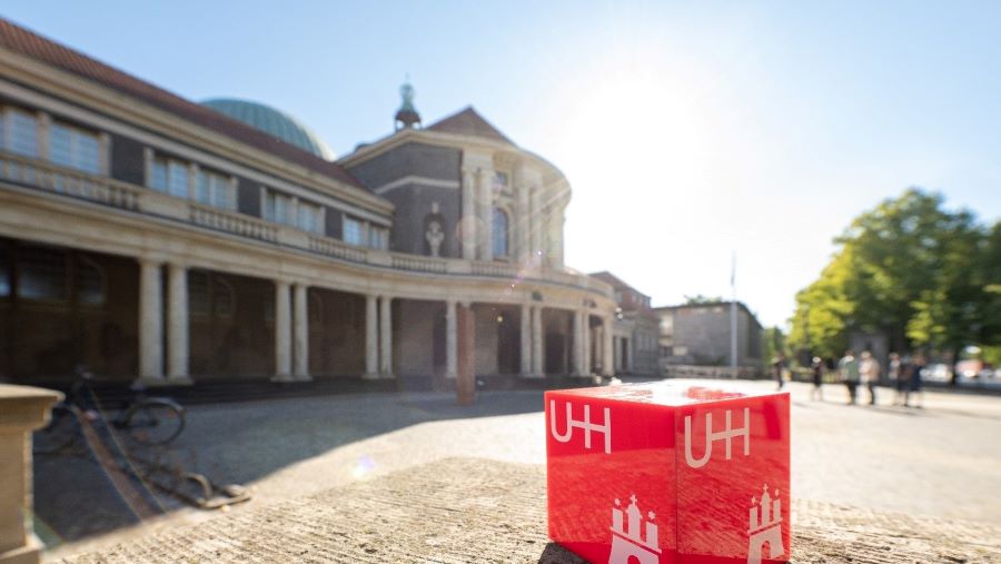 Das Foto zeigt das Hauptgebäude der Universität Hamburg. Im Vordergrund steht ein roter Würfel, auf dem das Logo der Universität Hamburg abgebildet ist.