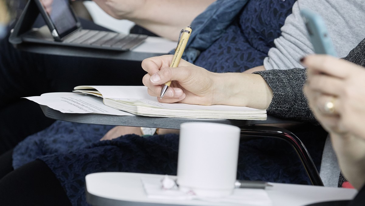In einer Reihe sitzen Personen, deren Hände schreiben oder ein Notebook bedienen.